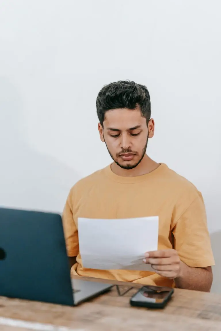 Serious young bearded Hispanic businessman in casual outfit reading contract while sitting at table with laptop in office